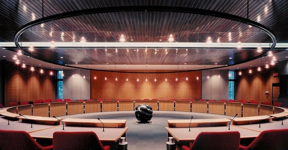 La salle du conseil de l'OACI à Montréal : un anneau de pupitres en bois, murs lambrissés, anneau de lumière au plafond